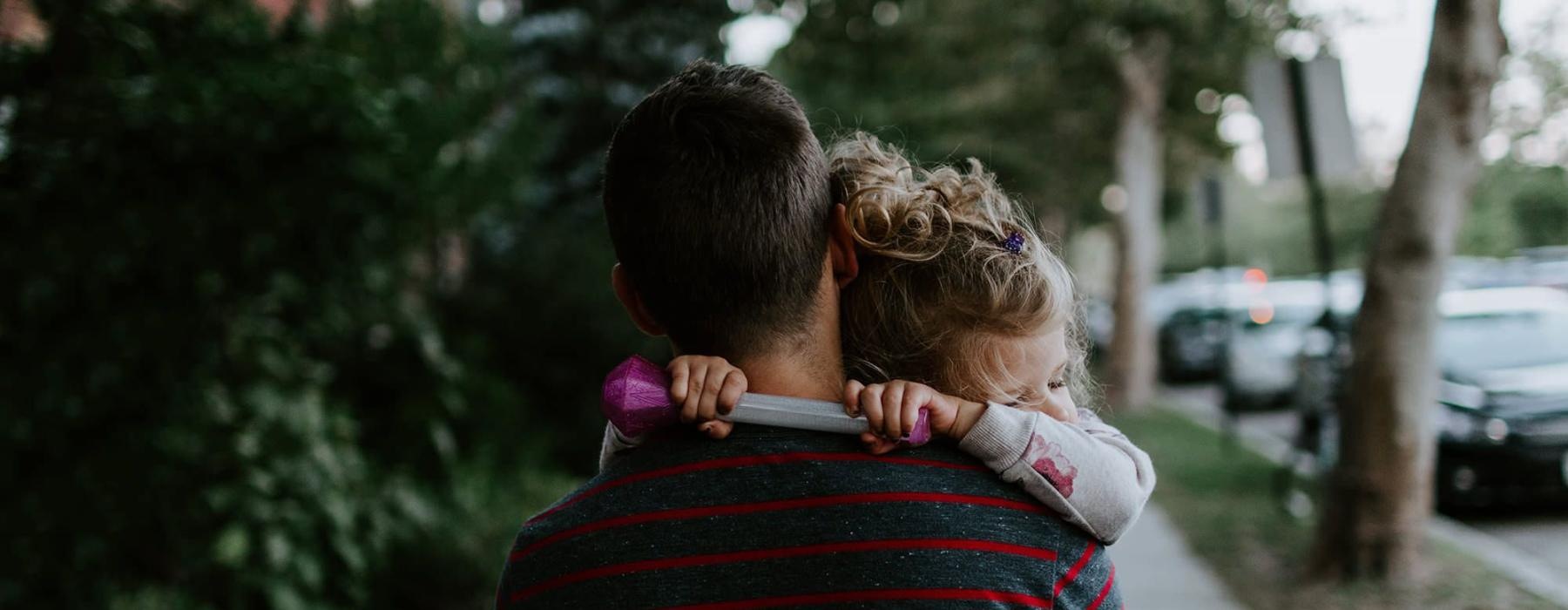 tired little girl rests her head on her father's shoulder as he carries her down the city sidewalk